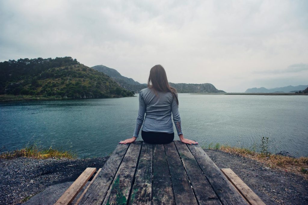 Femme portant une chemise grise à manches longues et un bas noir, assise sur une table de pique-nique en bois gris, face à une étendue d'eau calme, à l'heure du jour.